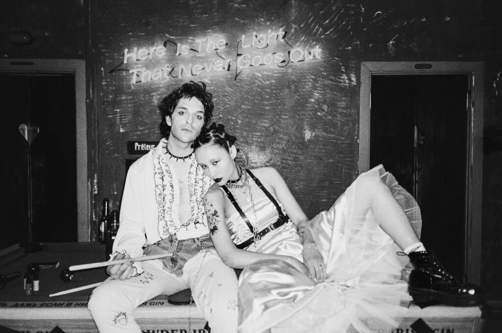 A stylish couple dressed in unique outfits, relaxing on a pool table in a moody room.